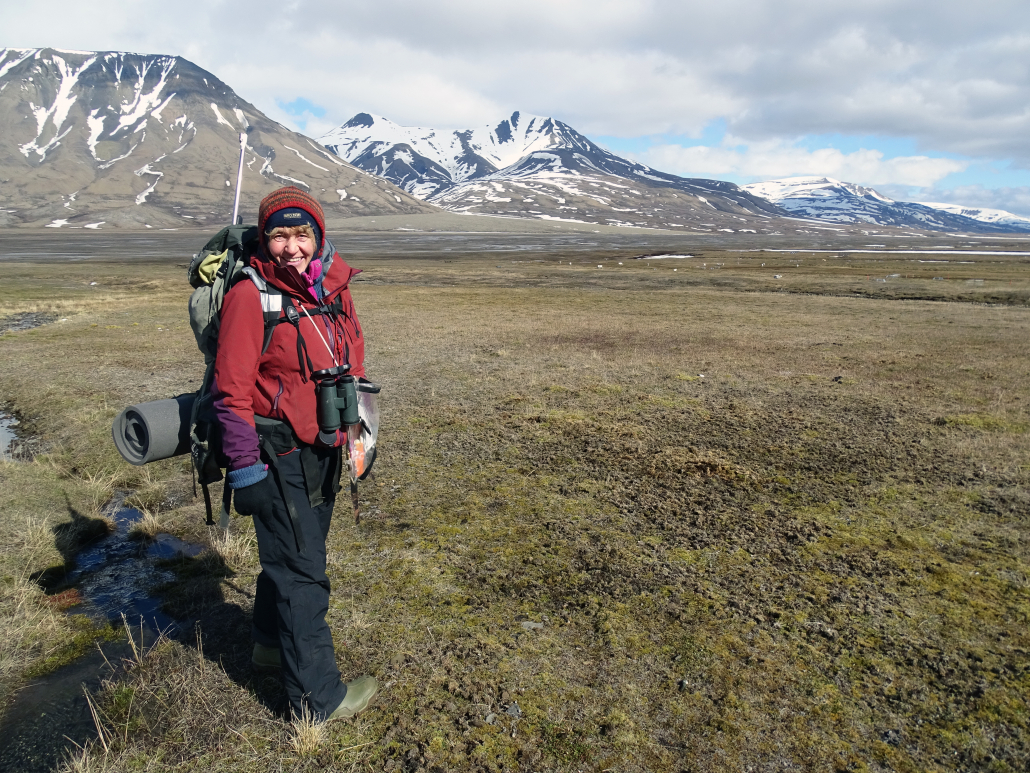 Kvinne står på marka i Svalbards natur, fjell og grønne sletter i omgivelsene 