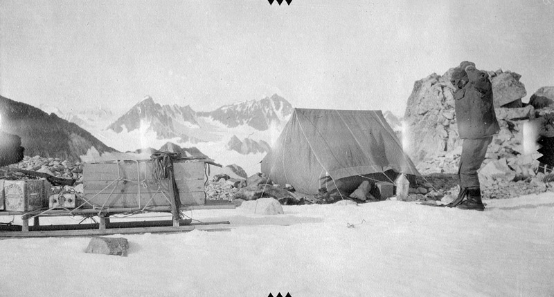 Mann står foran telt og slede, i bakgrunnen snedekket fjell