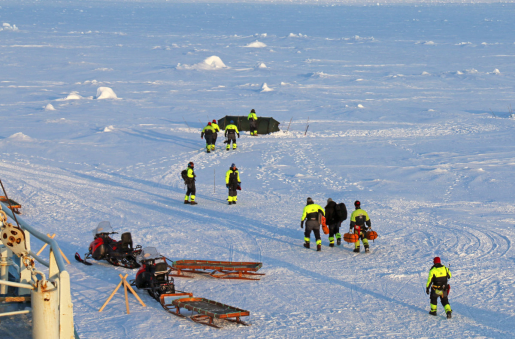 Nine people walking on the snow in survival suits, carrying equipment