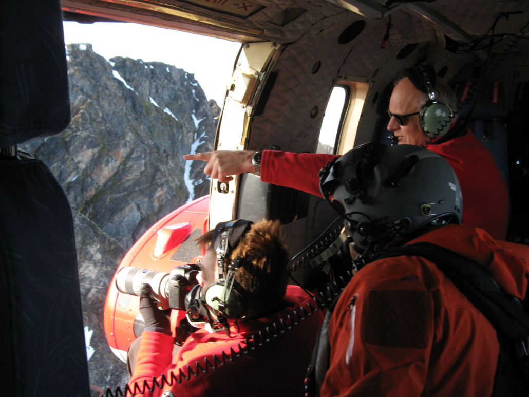 Tre personer står i åpen luke i helikopter i lufta