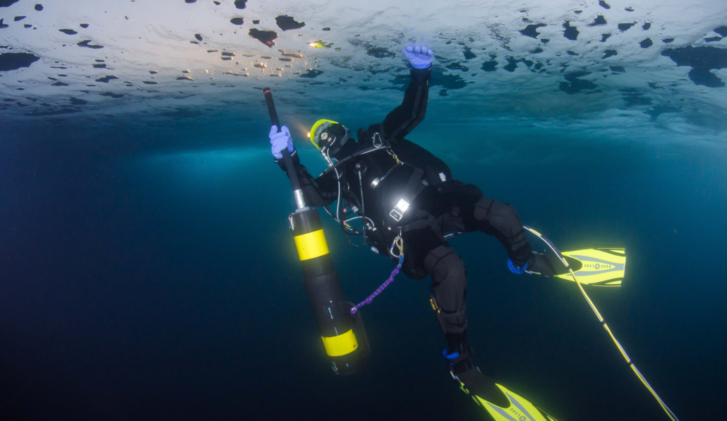 Person med dykkerdrakt i vannet holder et instrument i hånda under isen