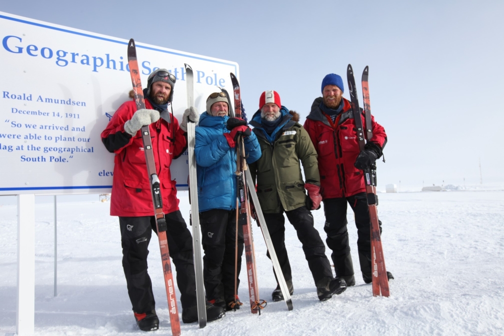 Fire menn poserer med ski og staver foran et skilt som markerer geografisk sørpol