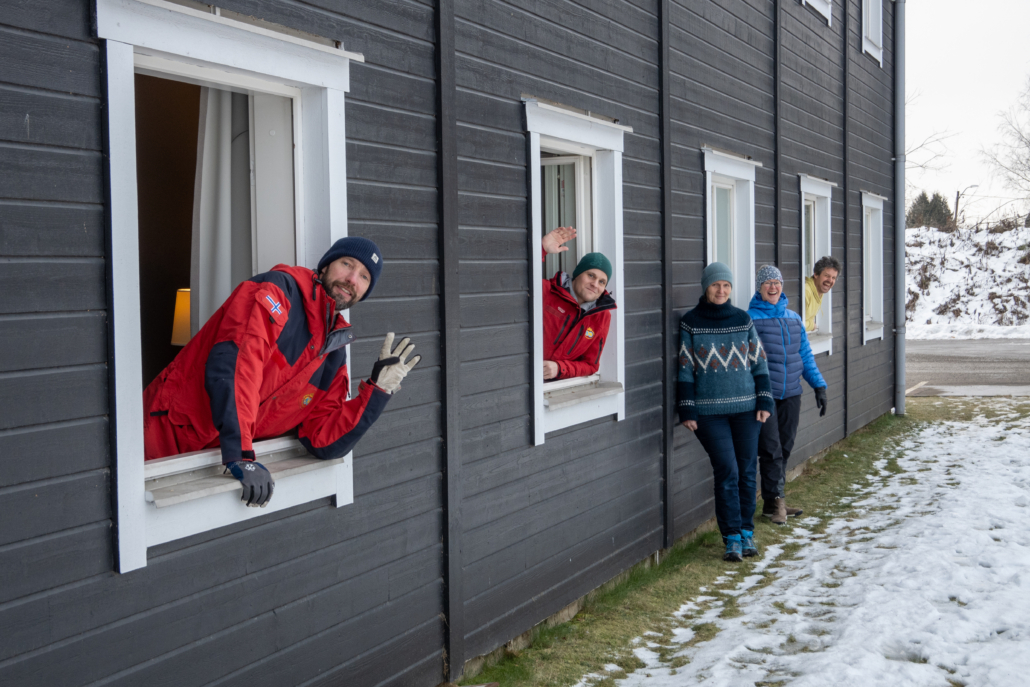 Tre personer stikker hodene ut av åpne vinduer og to personer står utenfor