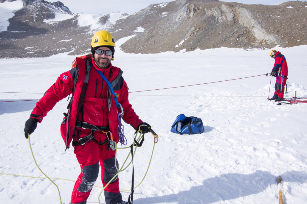 Person med røde klær og gul hjelm har tau i hendene, står på snø med fjell i bakgrunnen