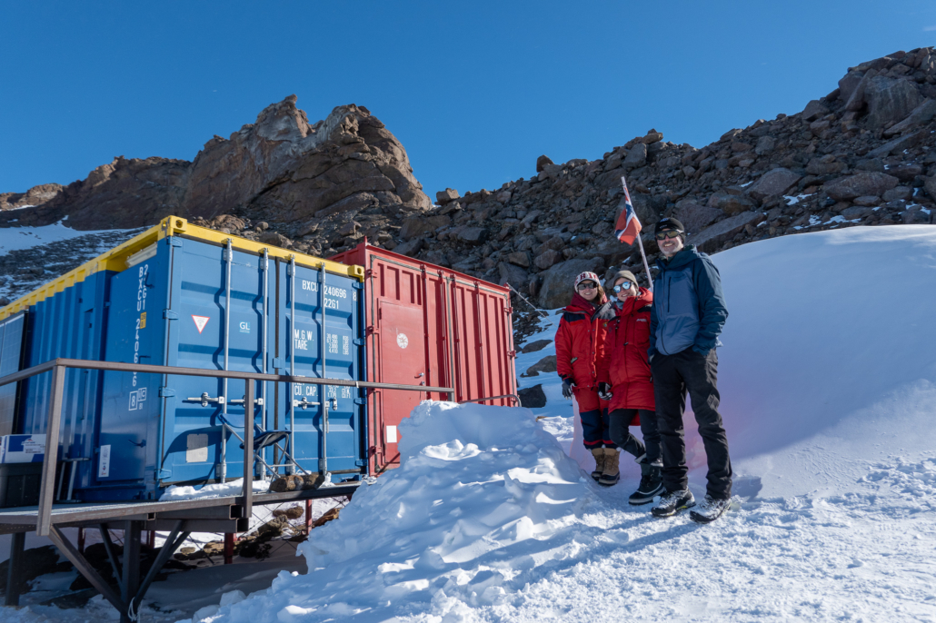 Tre personer står foran to kontainere med fjell i bakgrunnen