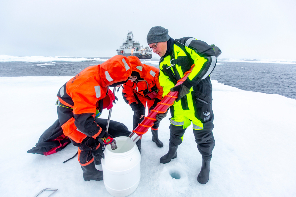 Tre personer med utstyr på isflak. Skip i bakgrunnen på havet