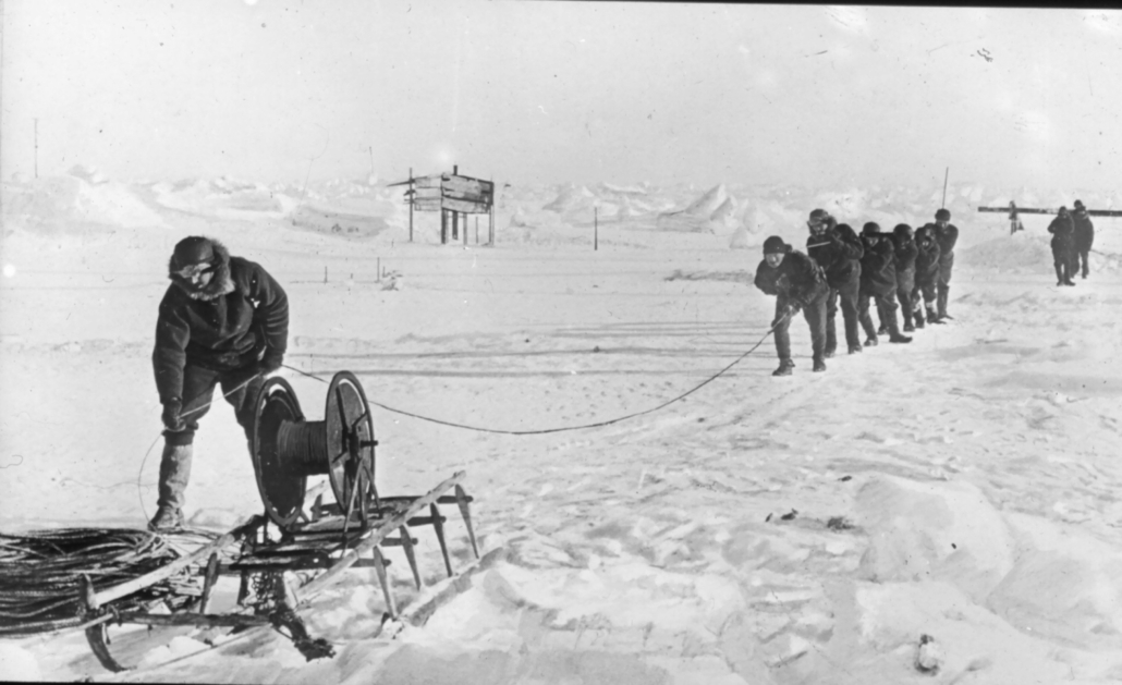 Svarthvitt bilde av menn på isen som drar opp en line fra et hull i isen
