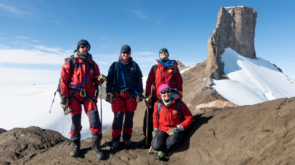 Fire personer står på steingrunn med fjelltopp i bakgrunnen