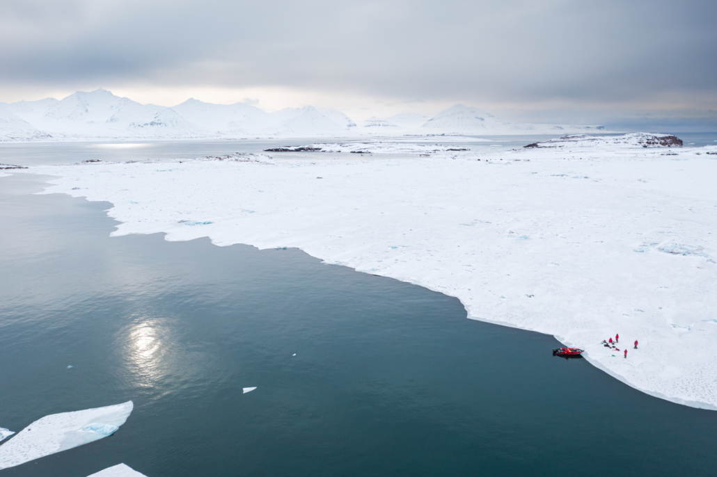 Landskap med hav og havis. En båt ligger fortøyd til isen og fire personer står på isen