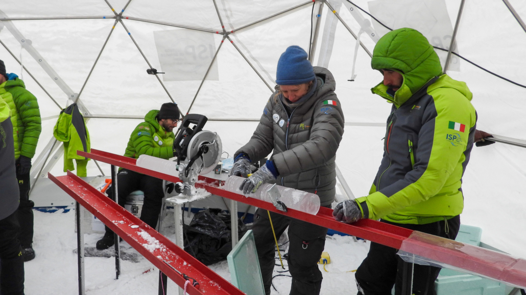 Tre personer med jakke, lue og hansker jobber med biter av is inne i et telt