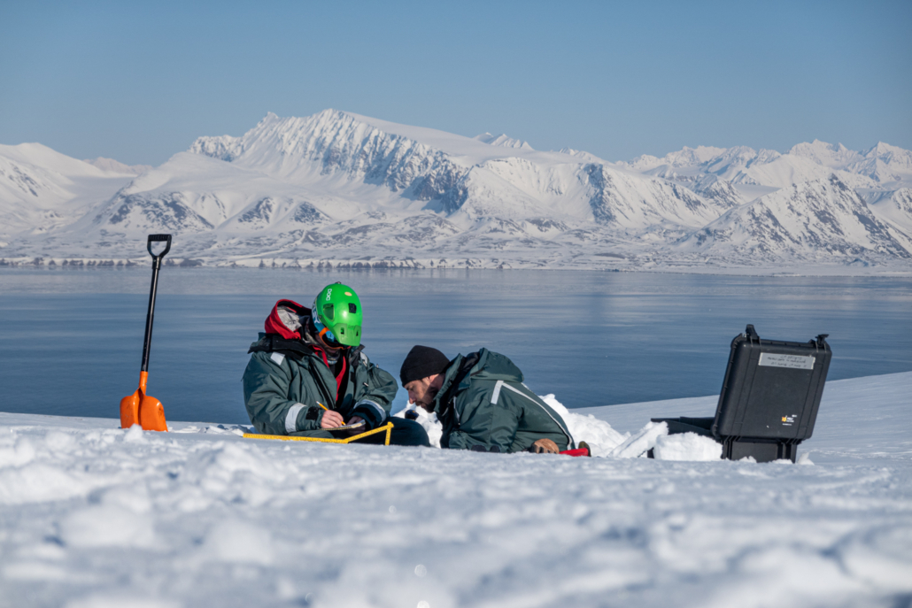 To personer sitter i snøen. Fjord med fjell i bakgrunnen