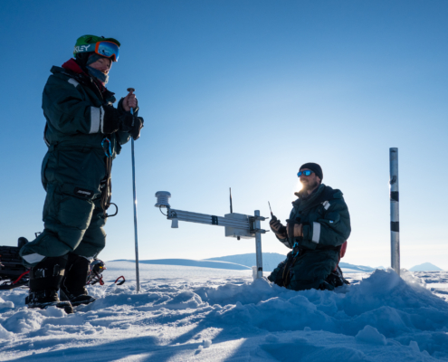 En person står og en person sitter på snø men de skruer på instrumenter