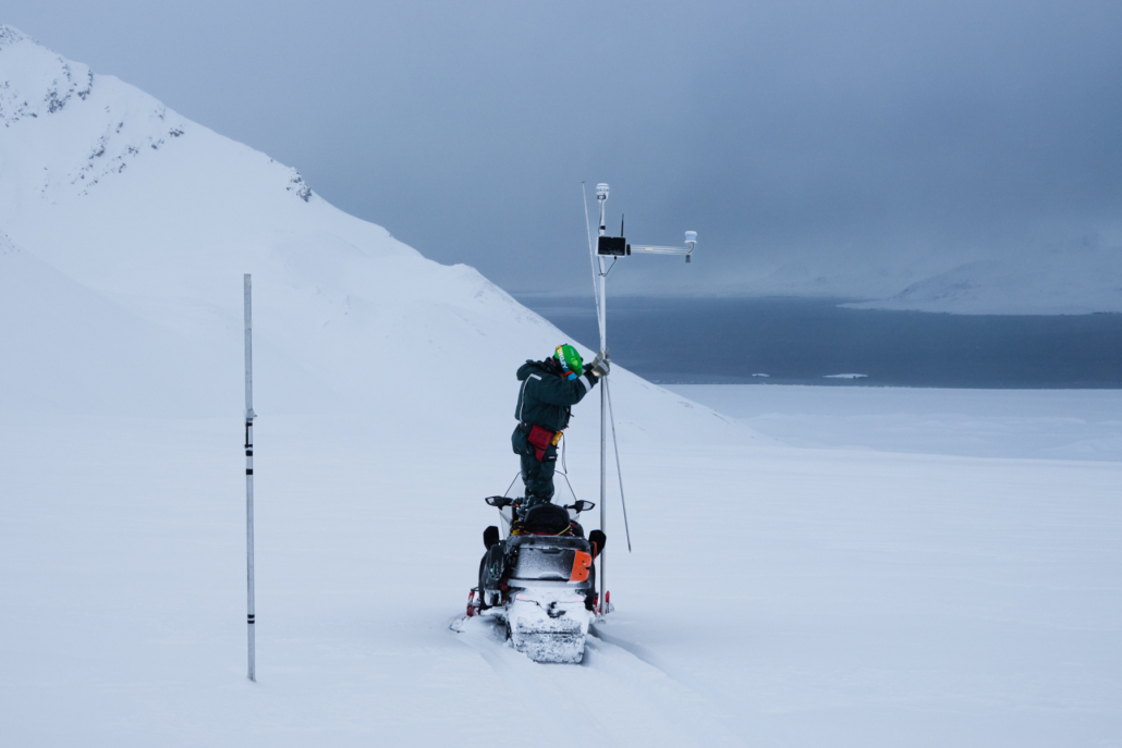 En person står på en snøskuter for å rekke opp til et instrument som står på en stang langt oppe