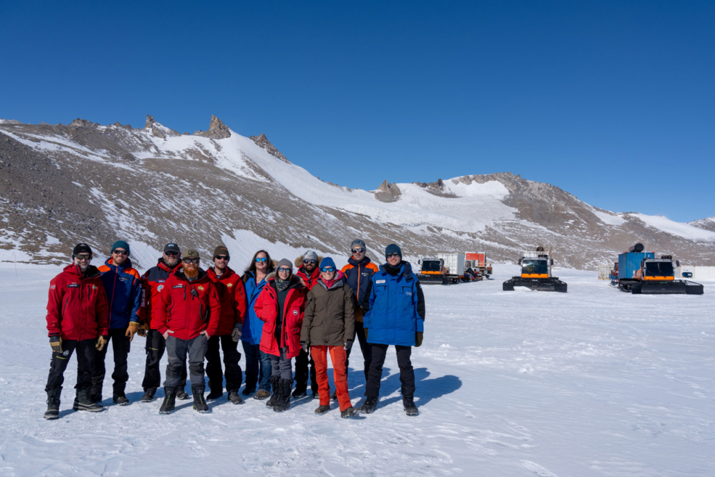En gruppe personer står på snø, i bakgrunnen er fjelltopper og tre kjøretøy